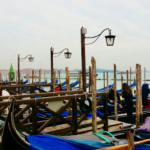 A line of traditional Venetian gondolas docked at wooden piers on a cloudy day, with black iron lamps overhead and the calm waters of the lagoon in the background. A must-see experience for any visitor looking for suggestions on what to do in Venice in 1 day, especially for those seeking a classic gondola ride.