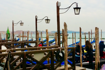 A line of traditional Venetian gondolas docked at wooden piers on a cloudy day, with black iron lamps overhead and the calm waters of the lagoon in the background. A must-see experience for any visitor looking for suggestions on what to do in Venice in 1 day, especially for those seeking a classic gondola ride.