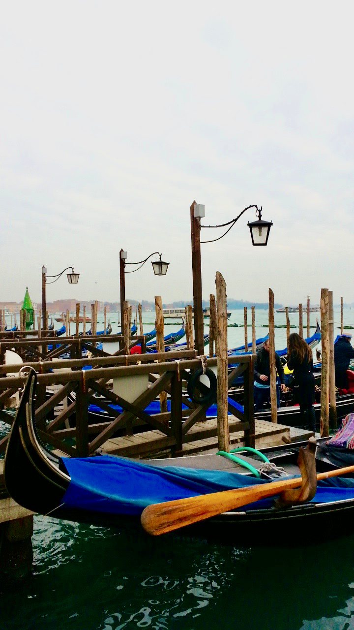 A line of traditional Venetian gondolas docked at wooden piers on a cloudy day, with black iron lamps overhead and the calm waters of the lagoon in the background. A must-see experience for any visitor looking for suggestions on what to do in Venice in 1 day, especially for those seeking a classic gondola ride.