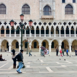Tourists walk and feed pigeons in front of the ornate facade of the Doge’s Palace in Piazza San Marco, Venice, Italy. The Gothic arches and intricate stonework highlight the city’s rich history, making this a top feature in any Venice Italy travel guide and a compelling reason when asking is it worth visiting Venice.