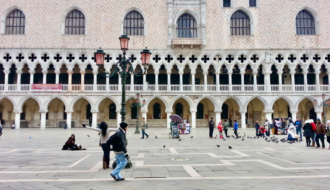 Tourists walk and feed pigeons in front of the ornate facade of the Doge’s Palace in Piazza San Marco, Venice, Italy. The Gothic arches and intricate stonework highlight the city’s rich history, making this a top feature in any Venice Italy travel guide and a compelling reason when asking is it worth visiting Venice.