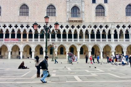 Tourists walk and feed pigeons in front of the ornate facade of the Doge’s Palace in Piazza San Marco, Venice, Italy. The Gothic arches and intricate stonework highlight the city’s rich history, making this a top feature in any Venice Italy travel guide and a compelling reason when asking is it worth visiting Venice.