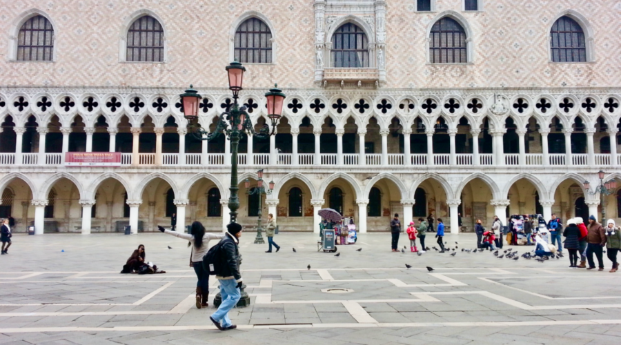 Tourists walk and feed pigeons in front of the ornate facade of the Doge’s Palace in Piazza San Marco, Venice, Italy. The Gothic arches and intricate stonework highlight the city’s rich history, making this a top feature in any Venice Italy travel guide and a compelling reason when asking is it worth visiting Venice.