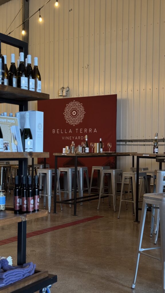 Interior of Bella Terra Vineyards in Niagara on the Lake featuring high top tasting tables, shelves of wine bottles, and a red wall sign that reads Bella Terra Vineyards. The modern rustic winery setting creates a relaxed and romantic wine tasting atmosphere perfect for valentine’s day in Canada.
