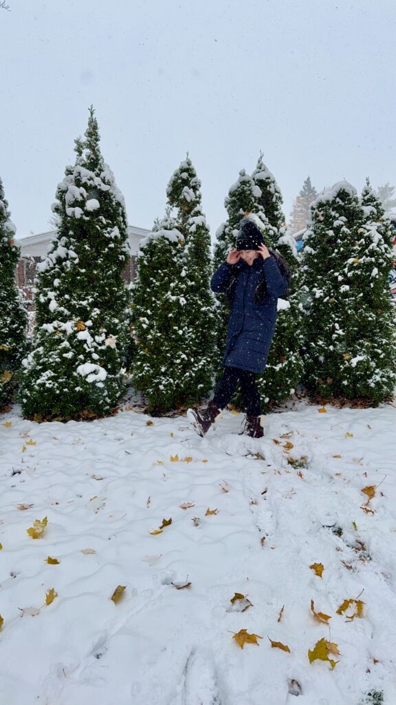 Woman walking through fresh snow in a long dark winter coat, knit hat, and lace up boots, surrounded by tall evergreen trees dusted in white. The cozy layered outfit and snowy backdrop capture practical winter packing outfits perfect for staying warm during valentine’s day in Canada.