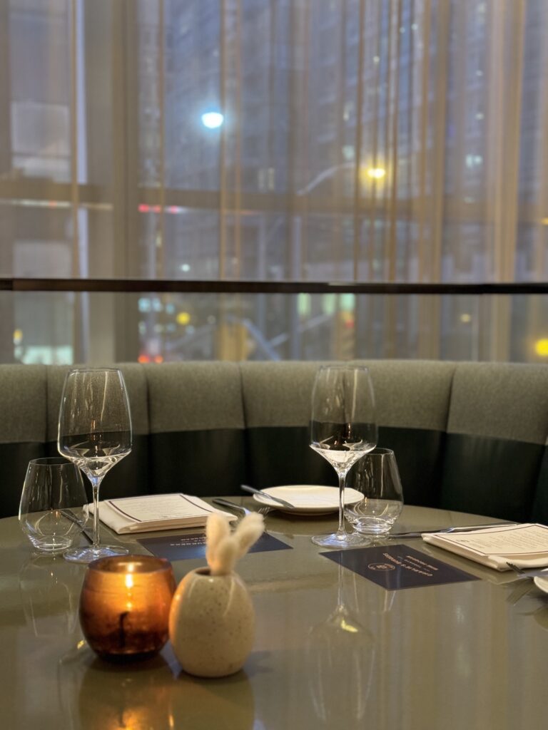 Candlelit table setting at Four Seasons Toronto featuring wine glasses, neatly folded napkins, and a curved velvet banquette overlooking the city at dusk. The intimate upscale ambiance creates a refined and romantic atmosphere for celebrating valentine’s day in Canada.