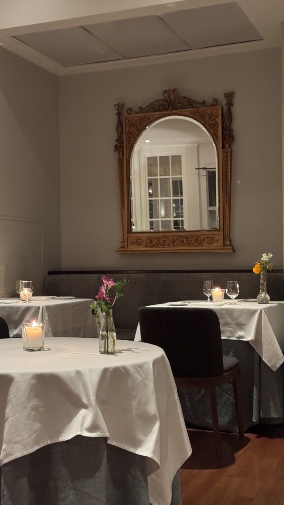 Elegant dining room at Langdon Hall in Cambridge featuring white tablecloth tables set with wine glasses, soft candlelight, and small floral arrangements beneath an ornate gold framed mirror. The intimate refined ambiance creates a romantic countryside setting perfect for celebrating valentine’s day in Canada.