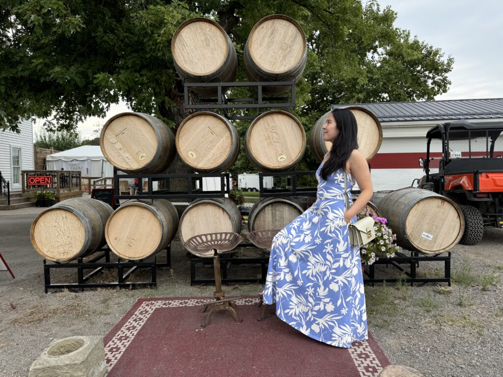 Woman in a blue floral maxi dress posing in front of stacked wooden wine barrels at Sue Ann Staff Winery in Niagara on the Lake. The charming vineyard setting and rustic barrel backdrop create a romantic wine country scene perfect for celebrating valentine’s day in Canada.