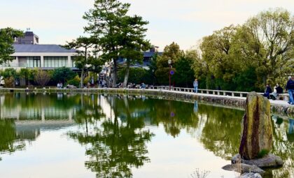 Peaceful pond in Nara surrounded by tall green trees and a walking path with visitors enjoying the scenery during a Nara day trip. The still water reflects the trees, stone walkway, and nearby buildings under a soft evening sky.