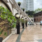 A polished courtyard at 1881 Heritage lined with trimmed potted shrubs and classic round lanterns, leading to escalators and surrounded by colonial and modern buildings. The wet pavement hints at recent rain, offering a quiet pause in one day in Hong Kong.