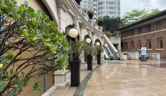 A polished courtyard at 1881 Heritage lined with trimmed potted shrubs and classic round lanterns, leading to escalators and surrounded by colonial and modern buildings. The wet pavement hints at recent rain, offering a quiet pause in one day in Hong Kong.