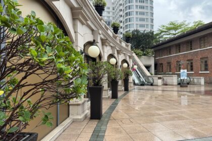 A polished courtyard at 1881 Heritage lined with trimmed potted shrubs and classic round lanterns, leading to escalators and surrounded by colonial and modern buildings. The wet pavement hints at recent rain, offering a quiet pause in one day in Hong Kong.