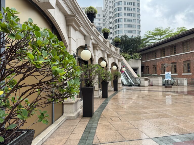 A polished courtyard at 1881 Heritage lined with trimmed potted shrubs and classic round lanterns, leading to escalators and surrounded by colonial and modern buildings. The wet pavement hints at recent rain, offering a quiet pause in one day in Hong Kong.