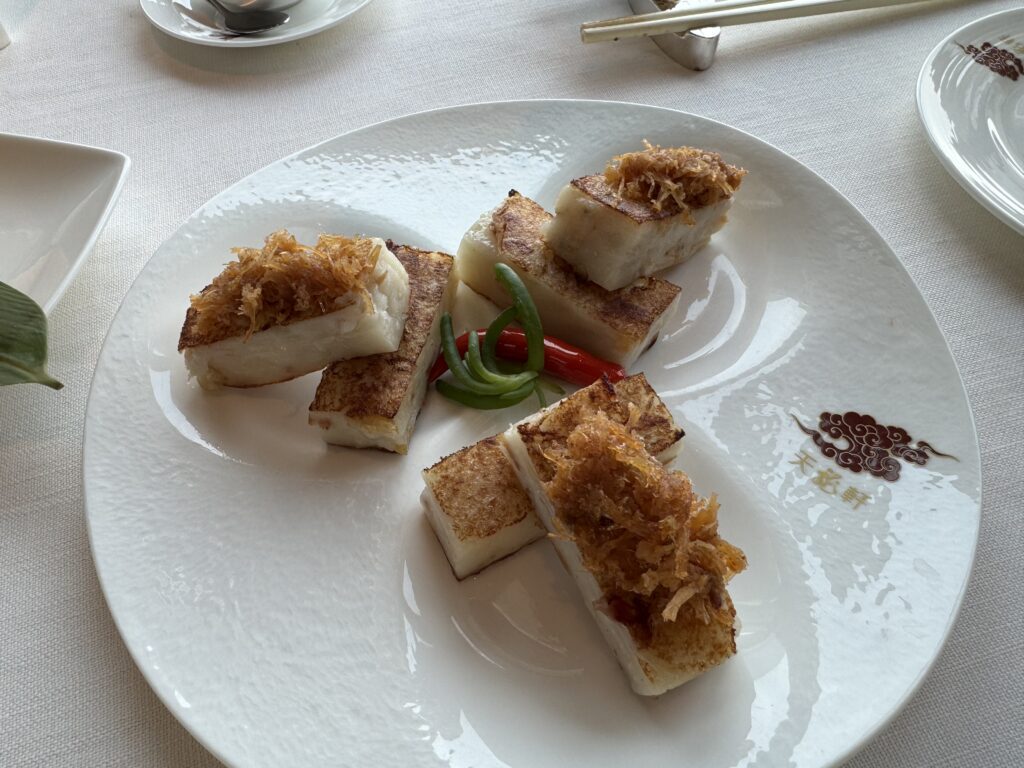 Elegant plate of pan-fried turnip cakes topped with crispy dried scallop threads, garnished with red and green chili slices, served at Tin Lung Heen in the Ritz-Carlton Hong Kong. A refined version of classic Hong Kong food presented with visual flair.