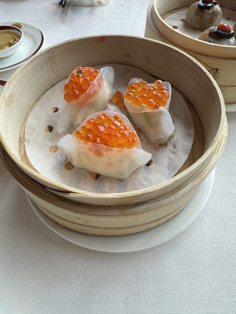 Bamboo steamer filled with translucent shrimp dumplings topped with vibrant orange salmon roe, served on parchment inside a dim sum restaurant. A modern twist on traditional Hong Kong food showcasing creative presentation.