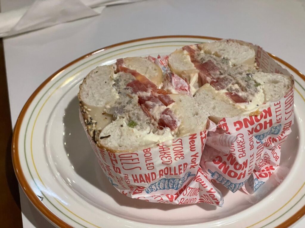 Cross-section of a loaded bagel sandwich wrapped in custom deli paper printed with “Hong Kong” and “New York,” filled with cream cheese, tomatoes, and fish spread. A fusion breakfast moment during one day in Hong Kong.