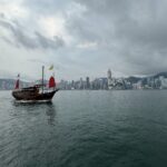 A traditional Chinese junk boat with red sails glides across Victoria Harbour under a cloudy sky, with the iconic Hong Kong skyline rising in the background. This scenic view captures the cultural and modern contrast perfect for a Hong Kong travel itinerary in 3 days.