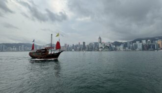 A traditional Chinese junk boat with red sails glides across Victoria Harbour under a cloudy sky, with the iconic Hong Kong skyline rising in the background. This scenic view captures the cultural and modern contrast perfect for a Hong Kong travel itinerary in 3 days.