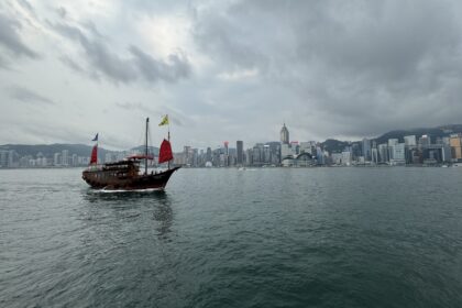 A traditional Chinese junk boat with red sails glides across Victoria Harbour under a cloudy sky, with the iconic Hong Kong skyline rising in the background. This scenic view captures the cultural and modern contrast perfect for a Hong Kong travel itinerary in 3 days.