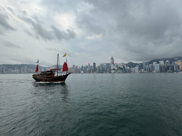 A traditional Chinese junk boat with red sails glides across Victoria Harbour under a cloudy sky, with the iconic Hong Kong skyline rising in the background. This scenic view captures the cultural and modern contrast perfect for a Hong Kong travel itinerary in 3 days.