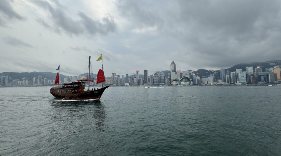 A traditional Chinese junk boat with red sails glides across Victoria Harbour under a cloudy sky, with the iconic Hong Kong skyline rising in the background. This scenic view captures the cultural and modern contrast perfect for a Hong Kong travel itinerary in 3 days.