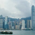 A harbour cruise boat sails across Victoria Harbour in Hong Kong, with the iconic skyline in the background, including the tall International Finance Centre (IFC) tower, the Bank of China building, and the Hong Kong Observation Wheel under cloudy skies.