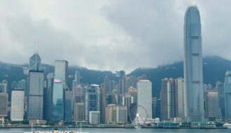 A harbour cruise boat sails across Victoria Harbour in Hong Kong, with the iconic skyline in the background, including the tall International Finance Centre (IFC) tower, the Bank of China building, and the Hong Kong Observation Wheel under cloudy skies.