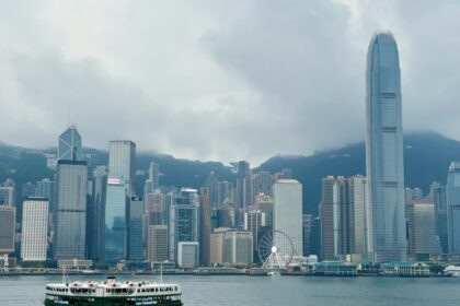 A harbour cruise boat sails across Victoria Harbour in Hong Kong, with the iconic skyline in the background, including the tall International Finance Centre (IFC) tower, the Bank of China building, and the Hong Kong Observation Wheel under cloudy skies.