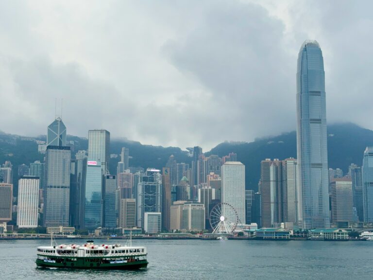 A harbour cruise boat sails across Victoria Harbour in Hong Kong, with the iconic skyline in the background, including the tall International Finance Centre (IFC) tower, the Bank of China building, and the Hong Kong Observation Wheel under cloudy skies.