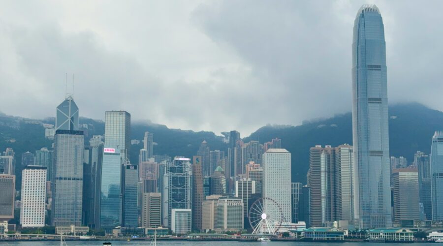 A harbour cruise boat sails across Victoria Harbour in Hong Kong, with the iconic skyline in the background, including the tall International Finance Centre (IFC) tower, the Bank of China building, and the Hong Kong Observation Wheel under cloudy skies.