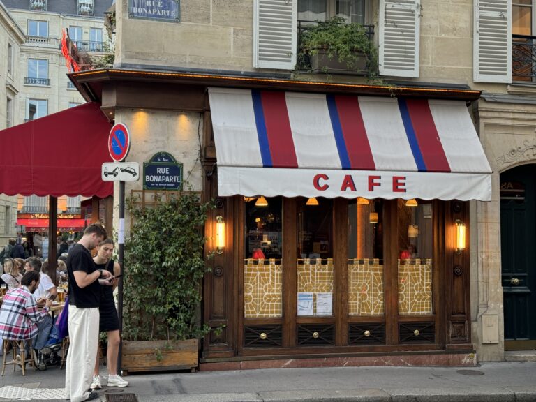 Street view of a Parisian café with a red, white, and blue striped awning labeled 'Café.' The exterior features wooden details, warm yellow lighting, and decorative patterns on the lower window panels. A green plant climbs along the wall under a sign for 'Rue Bonaparte.' Two people stand in the foreground looking at a phone, and outdoor seating with people dining is visible on the left.