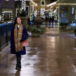 A woman in a navy blue coat, plaid scarf, and black boots walks confidently through a beautifully lit shopping district on a winter evening. She carries a pink purse and smiles as she enjoys the peaceful atmosphere, surrounded by festive string lights reflecting off the wet pavement. Overcoming pre-travel anxiety and travel-related stress, she looks relaxed and ready for her next adventure. Mastering pre-travel jitters can turn your trip into a walk in the park—just like this moment of calm before an exciting journey.