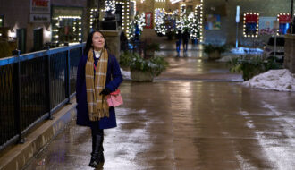 A woman in a navy blue coat, plaid scarf, and black boots walks confidently through a beautifully lit shopping district on a winter evening. She carries a pink purse and smiles as she enjoys the peaceful atmosphere, surrounded by festive string lights reflecting off the wet pavement. Overcoming pre-travel anxiety and travel-related stress, she looks relaxed and ready for her next adventure. Mastering pre-travel jitters can turn your trip into a walk in the park—just like this moment of calm before an exciting journey.