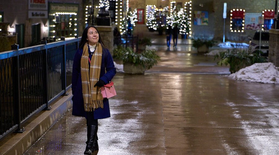 A woman in a navy blue coat, plaid scarf, and black boots walks confidently through a beautifully lit shopping district on a winter evening. She carries a pink purse and smiles as she enjoys the peaceful atmosphere, surrounded by festive string lights reflecting off the wet pavement. Overcoming pre-travel anxiety and travel-related stress, she looks relaxed and ready for her next adventure. Mastering pre-travel jitters can turn your trip into a walk in the park—just like this moment of calm before an exciting journey.