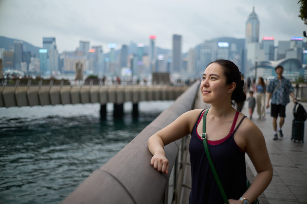 A woman in a blue dress stands on the boardwalk with views of Victoria Harbour in Hong Kong, with the iconic skyline in the background, including the tall International Finance Centre (IFC) tower, the Bank of China building, and the Hong Kong Observation Wheel under cloudy skies.”