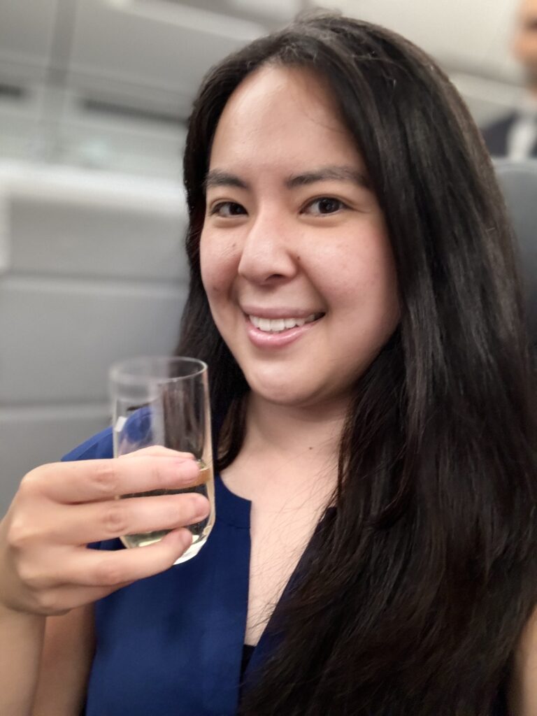 Woman holding a glass of white wine and smiling at the camera. She is on board Air France and heading towards Paris, France. She has a travel beauty routine she follows in flight and at the destination.