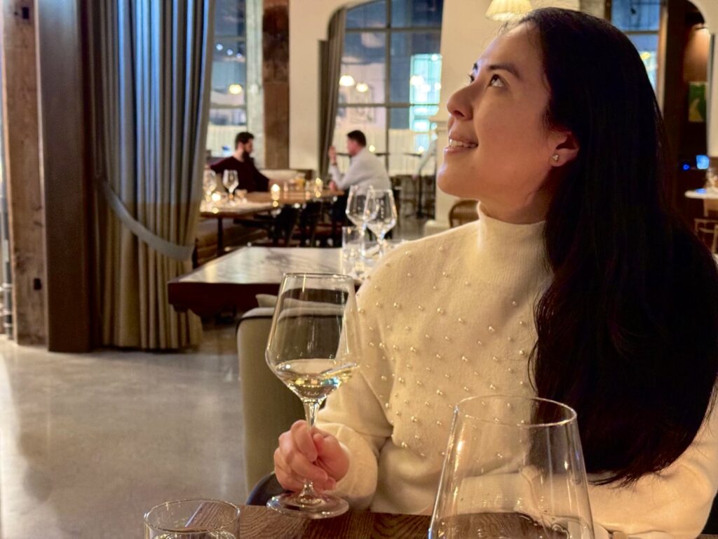 Woman looking up at the ceiling while holding a glass of white wine inside a restaurant with modern decor. She has a travel beauty routine to keep her skin glowing while travelling.