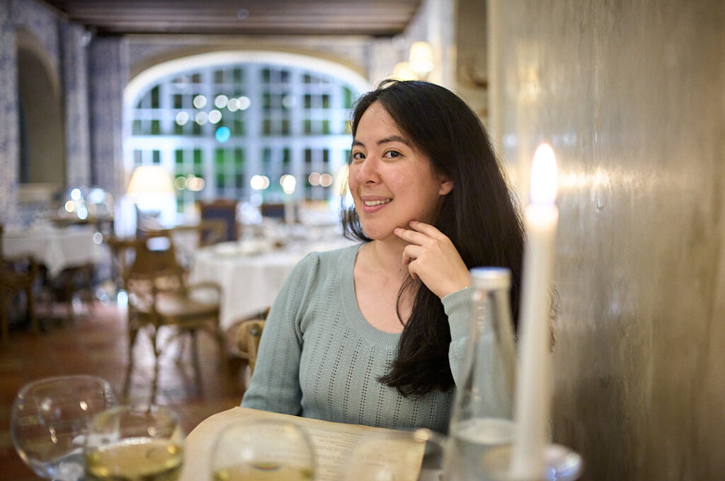 Woman smiles into camera inside a restaurant in Paris, France. There are glasses of wine on the table and a lit candle in the corner of the photograph.