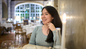 Woman smiles into camera inside a restaurant in Paris, France. There are glasses of wine on the table and a lit candle in the corner of the photograph.