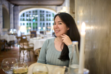 Woman smiles into camera inside a restaurant in Paris, France. There are glasses of wine on the table and a lit candle in the corner of the photograph.