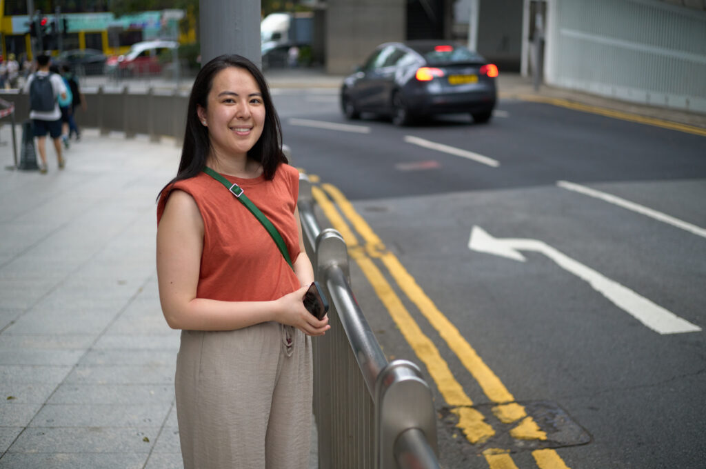 Woman smiling into camera wearing an orange shirt and brown pants with a green purse and she is holding a phone. She is in Hong Kong.