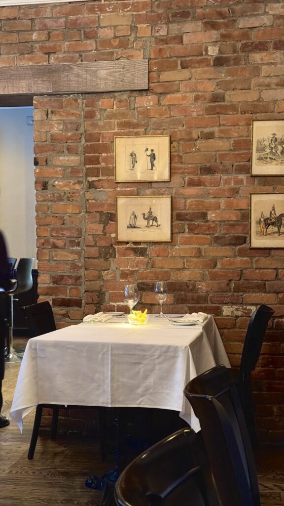 A romantic corner table set for two at Bonaparte Restaurant in Montreal, featuring a white tablecloth, wine glasses, and a glowing candle against an exposed brick wall with vintage framed art, offering a refined and intimate dining spot for a long weekend in Montreal. The warm tones and classic decor evoke timeless charm and elegance.