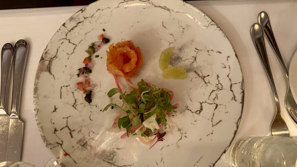 Alt text: A gourmet appetizer plated with artistic precision features a rose-shaped smoked salmon, citrus segments, microgreens, and a small dice of pickled vegetables on a marbled white dish. This elegant presentation reflects the fine dining experience found at upscale places to eat in Montreal.