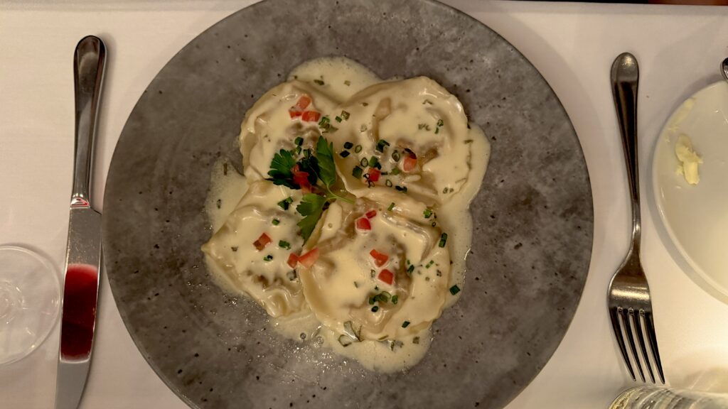 Alt text: A plate of handmade ravioli covered in a creamy white sauce is garnished with chopped chives, diced red pepper, and a sprig of parsley, served on a dark stoneware plate. This elegant pasta dish showcases the refined Italian cuisine available at upscale places to eat in Montreal.