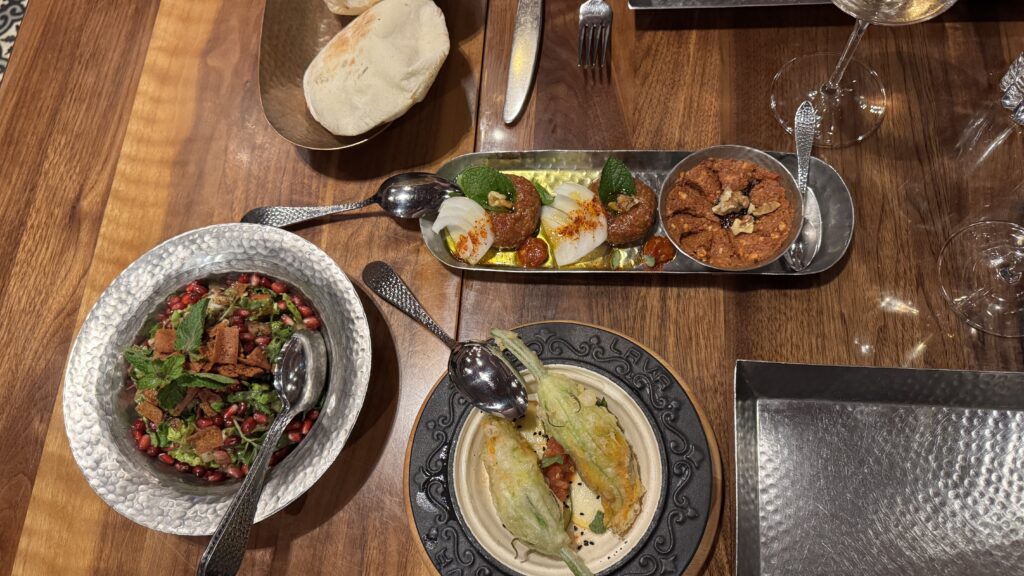 An elegant spread of Syrian mezze at Damas in Montreal, featuring vibrant dishes like fattoush with pomegranate, muhammara, kibbeh, and stuffed squash blossoms, paired with warm pita and a glass of wine—an indulgent and flavourful highlight of a long weekend in Montreal. Additional plates include kataifi-wrapped shrimp on creamy sauce and prosciutto-wrapped bites topped with quail egg, all set against the restaurant’s richly decorated interior.