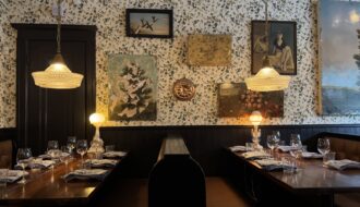A cozy interior of Joe Beef restaurant in Montreal, with two wooden booths set for dinner under soft pendant lighting and surrounded by vintage art and floral wallpaper, perfect for a memorable meal during a long weekend in Montreal. The eclectic decor and warm glow create an inviting, intimate dining atmosphere.