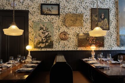 A cozy interior of Joe Beef restaurant in Montreal, with two wooden booths set for dinner under soft pendant lighting and surrounded by vintage art and floral wallpaper, perfect for a memorable meal during a long weekend in Montreal. The eclectic decor and warm glow create an inviting, intimate dining atmosphere.