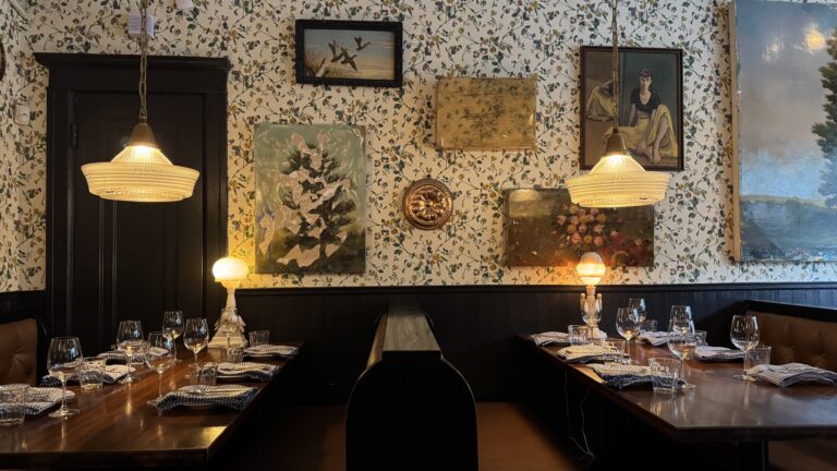 A cozy interior of Joe Beef restaurant in Montreal, with two wooden booths set for dinner under soft pendant lighting and surrounded by vintage art and floral wallpaper, perfect for a memorable meal during a long weekend in Montreal. The eclectic decor and warm glow create an inviting, intimate dining atmosphere.