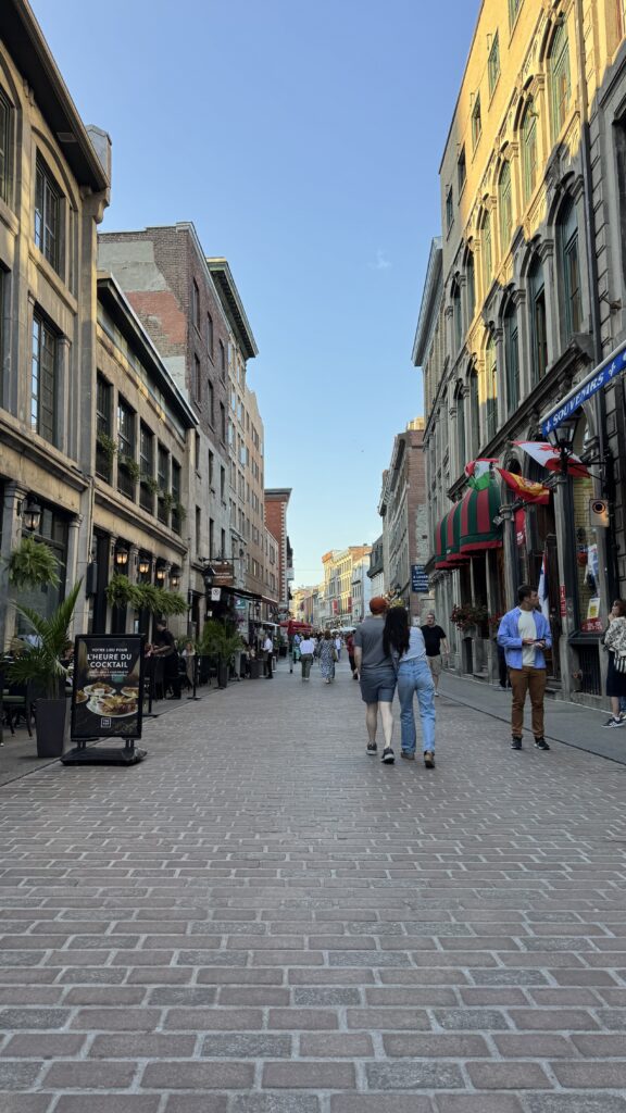 A cobblestone street in Old Montreal filled with people strolling under a clear blue sky, surrounded by historic buildings, cafés with patio seating, and souvenir shops, capturing a vibrant summer vibe perfect for a long weekend in Montreal.