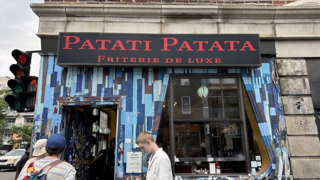 The colourful, patchwork-painted exterior of Patati Patata Friterie de Luxe in Montreal, with its bold red signage and cozy corner location, makes it a quirky and beloved spot for casual eats during a long weekend in Montreal. A small line of people waits to order under the soft glow of retro-style pendant lights.